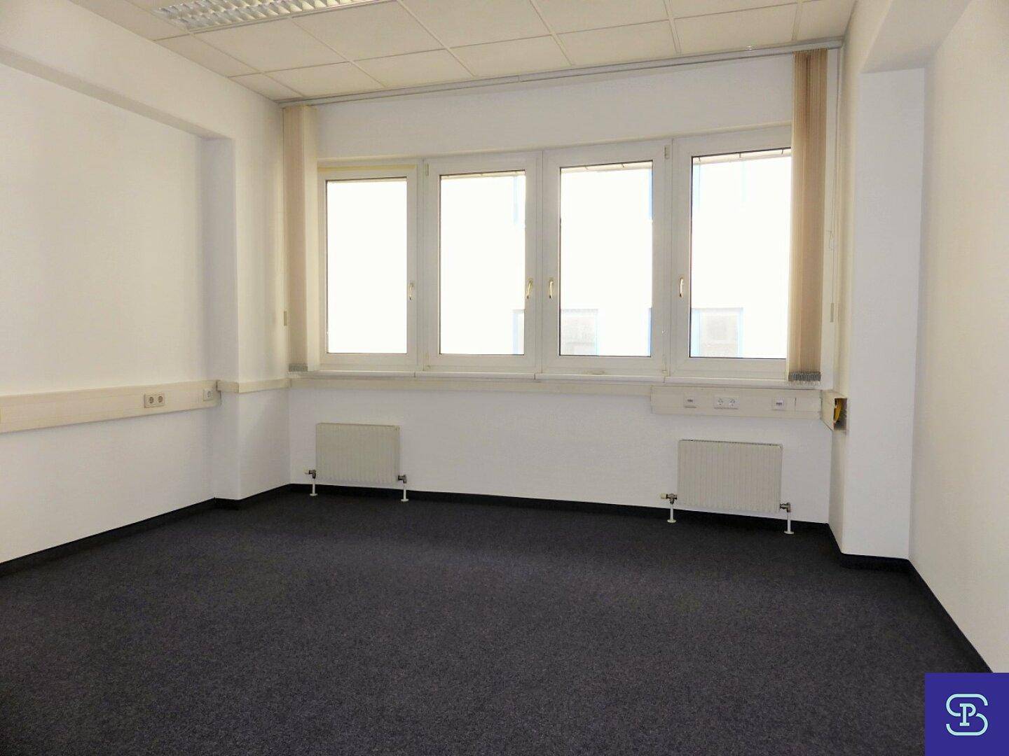 Büro mit breiter Fensterfront, die viel Licht hereinlässt, Heizkörpern und dunklem Teppichboden.