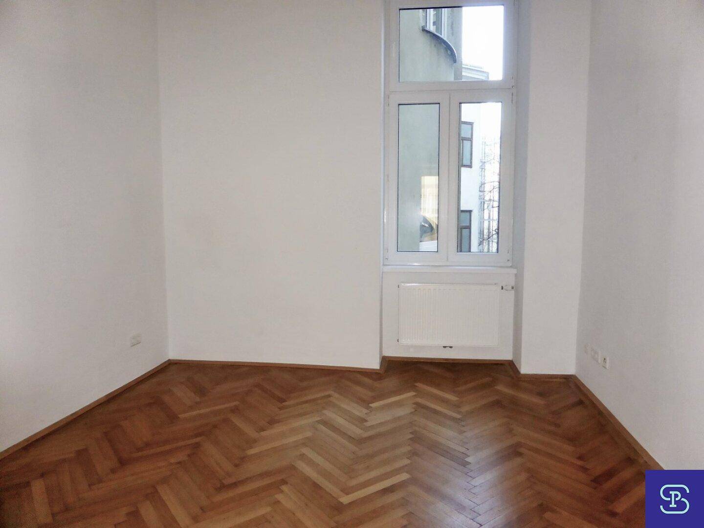 Heller Raum mit Fischgrätparkett und einem hohen Fenster, das viel Tageslicht spendet.