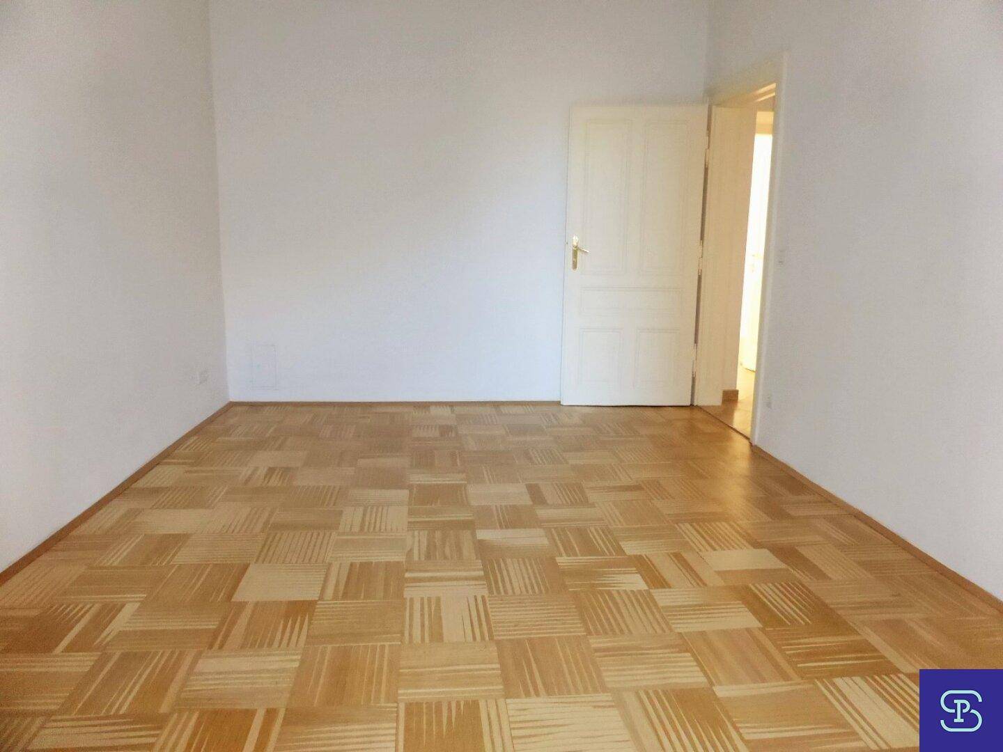 Geräumiges Zimmer mit quadratischem Parkettboden und einer weißen Tür, die zu einem anderen Raum führt.