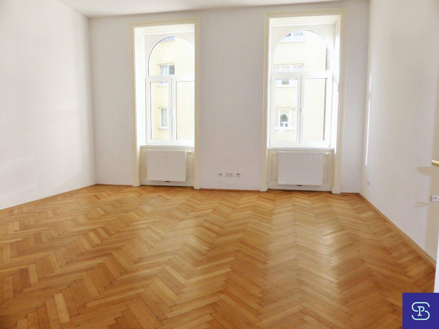Heller Raum mit Fischgrätparkett und zwei hohen Bogenfenstern, die den Blick nach draußen freigeben.