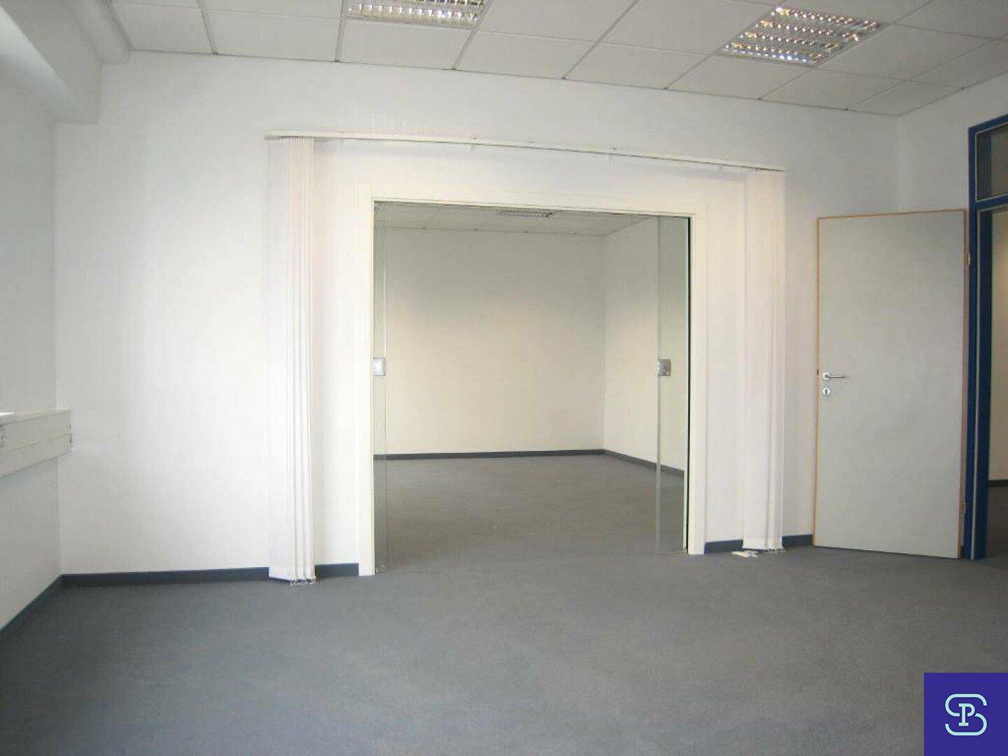 Leeres Büro mit Teppichboden und einer Glastür, die zu einem weiteren Raum führt.