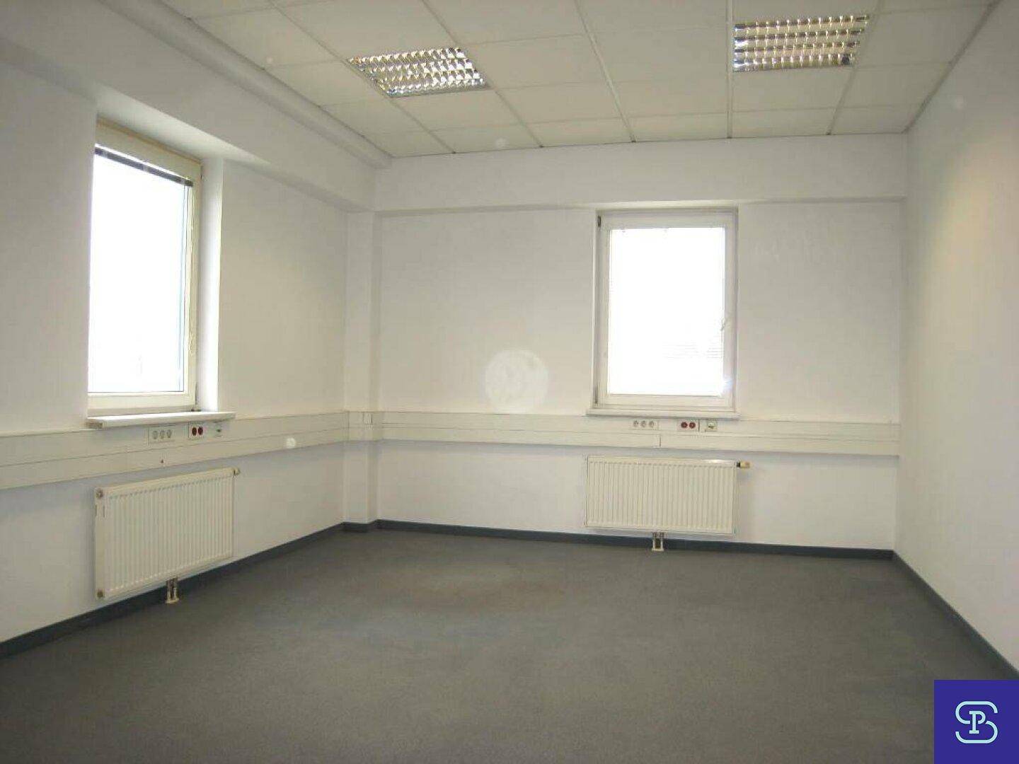 Eckbüro mit Teppichboden und zwei Fenstern, die für reichlich Tageslicht und eine offene Atmosphäre sorgen.