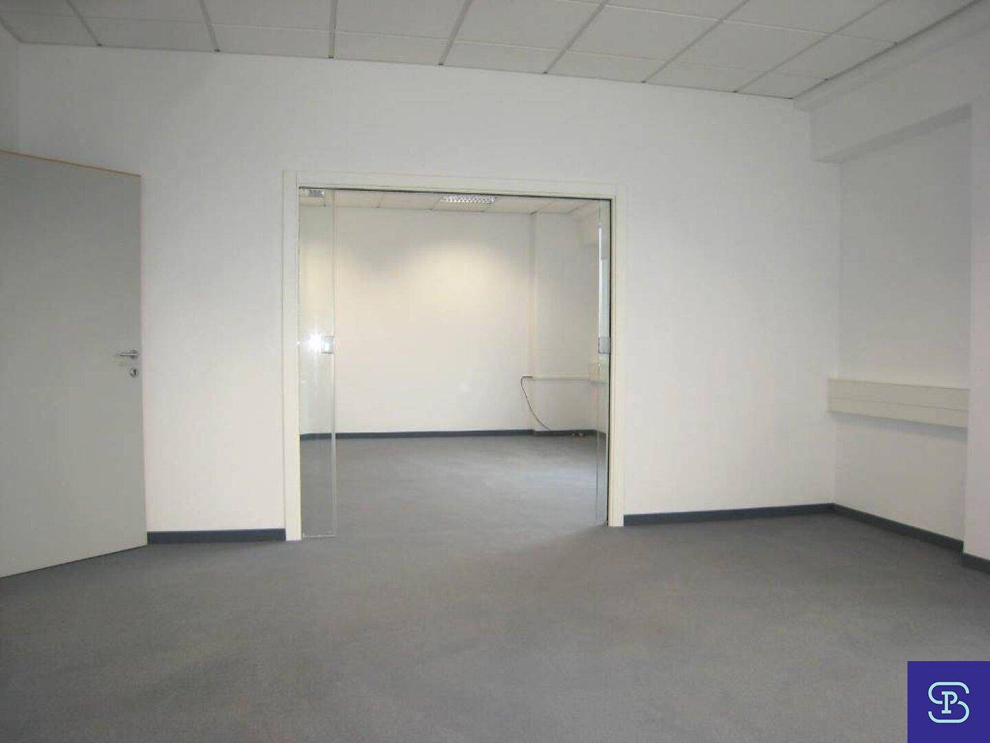 Büro mit Teppichboden und einer breiten Glas-Schiebetür, die einen offenen Übergang zu einem Nebenraum schafft.