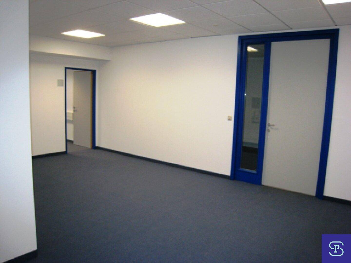 Leerer Büroraum mit blauen Türrahmen und Teppichboden, der Zugang zu weiteren Bereichen bietet.