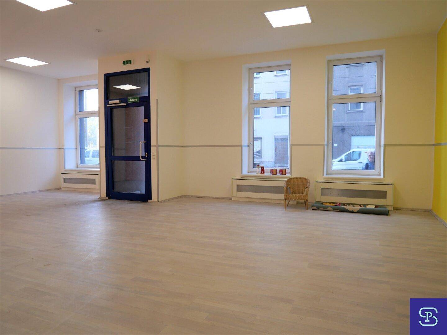 Geräumiger, heller Mehrzweckraum mit großen Fenstern, hellem Laminatboden und Blick auf die Straße.