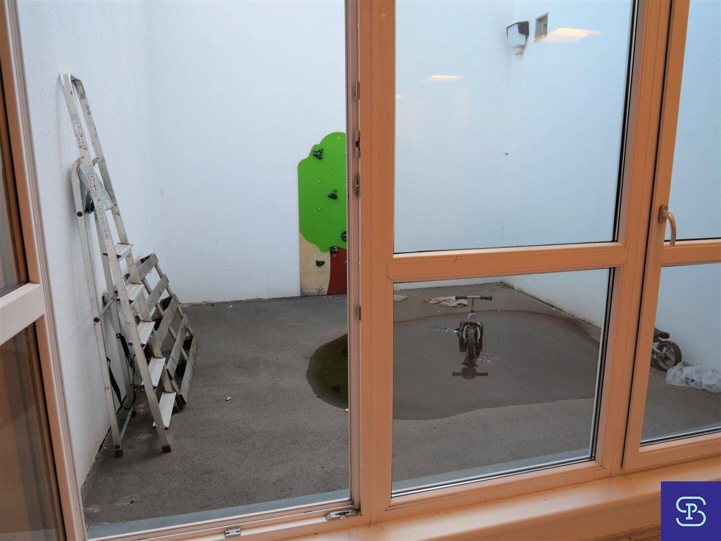 Blick auf einen kleinen, ummauerten Innenhof mit Betonboden, einer Leiter und Kinderspielgeräten, der Renovierung benötigt.