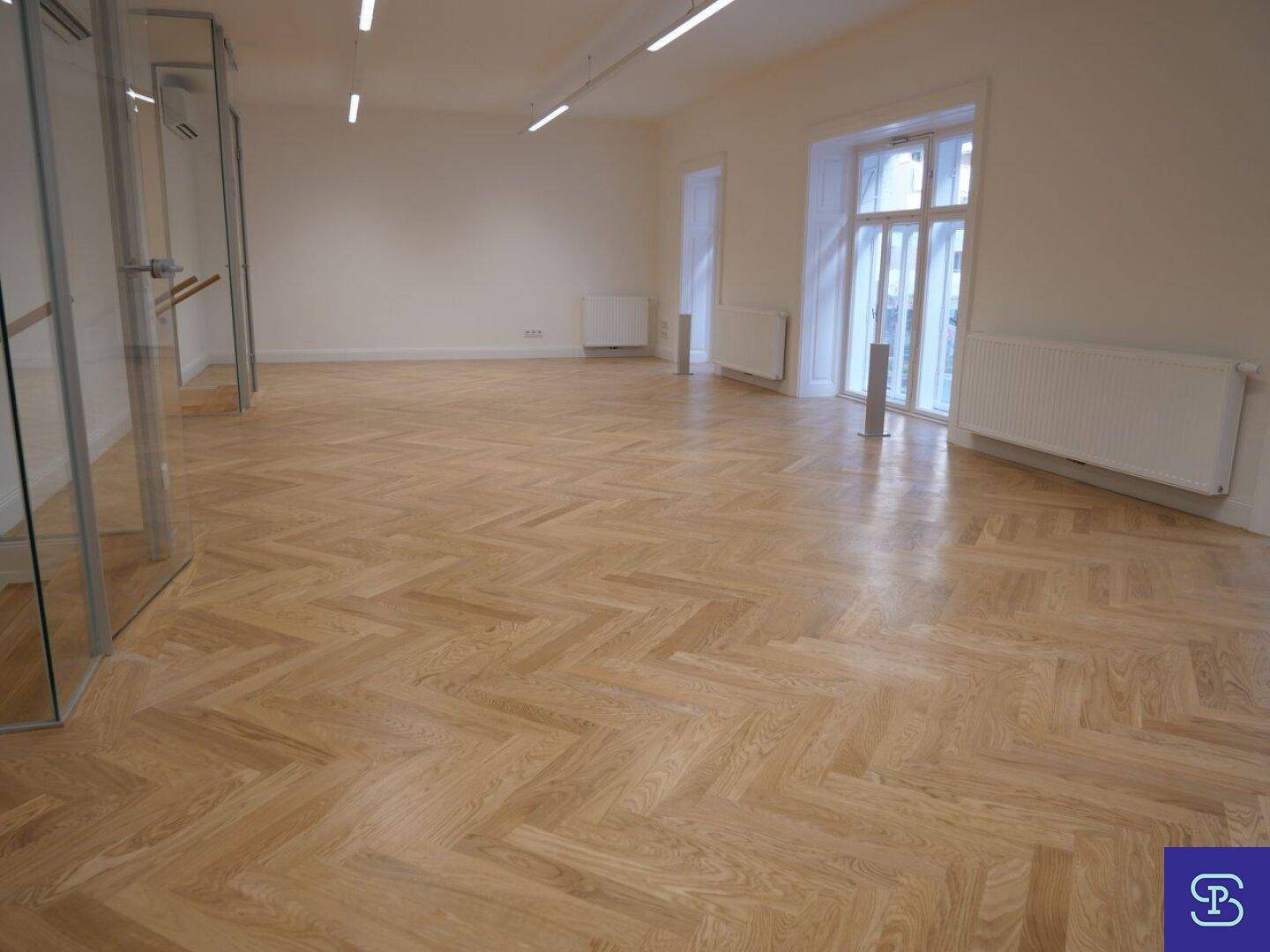 Weitläufiger Raum mit elegantem Fischgrätparkett und viel Tageslicht durch hohe Fenster.