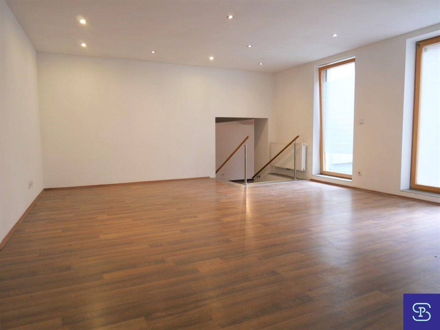 Geräumiger, unmöblierter Raum mit Laminatboden, Einbaustrahlern und Zugang zu einem Treppenhaus.