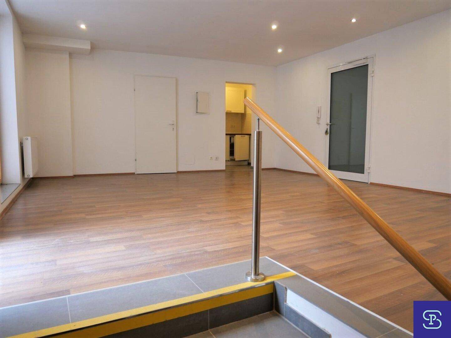 Großer, heller Raum mit Laminatboden, Treppenaufgang und Blick auf eine kleine Küchenzeile.