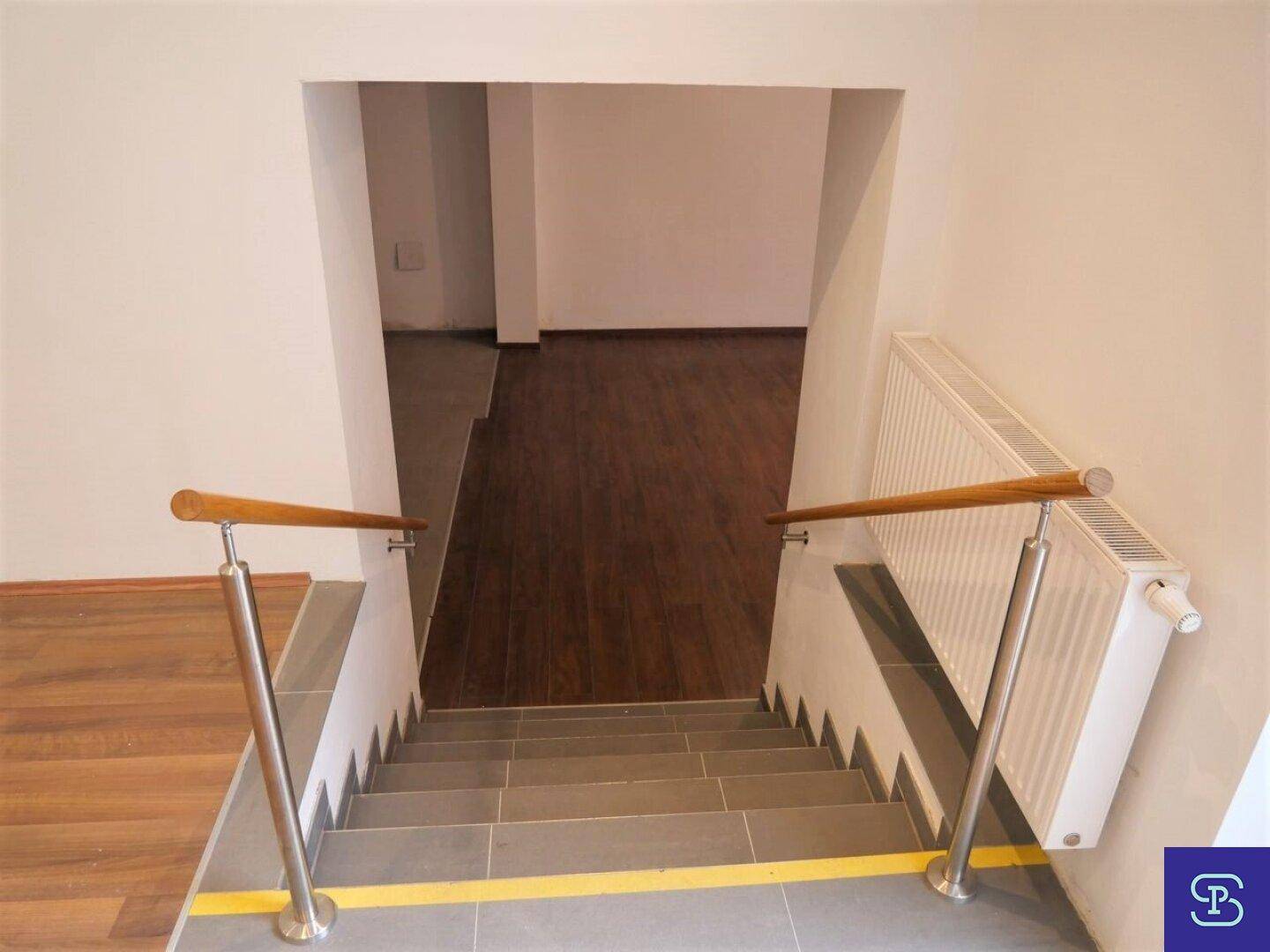 Treppenhaus mit Holzgeländer, Edelstahlpfosten und Fliesenboden, führt in einen unteren Bereich.