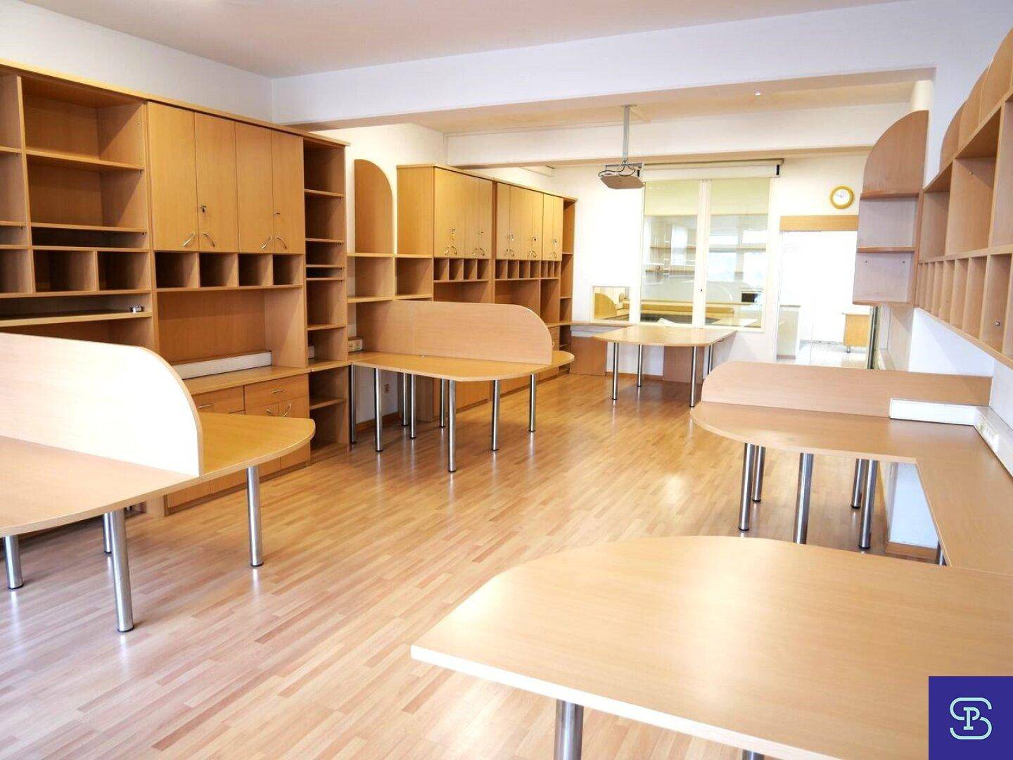 Heller Büroraum mit mehreren Schreibtischen, Holzregalen und einem Beamer an der Decke.