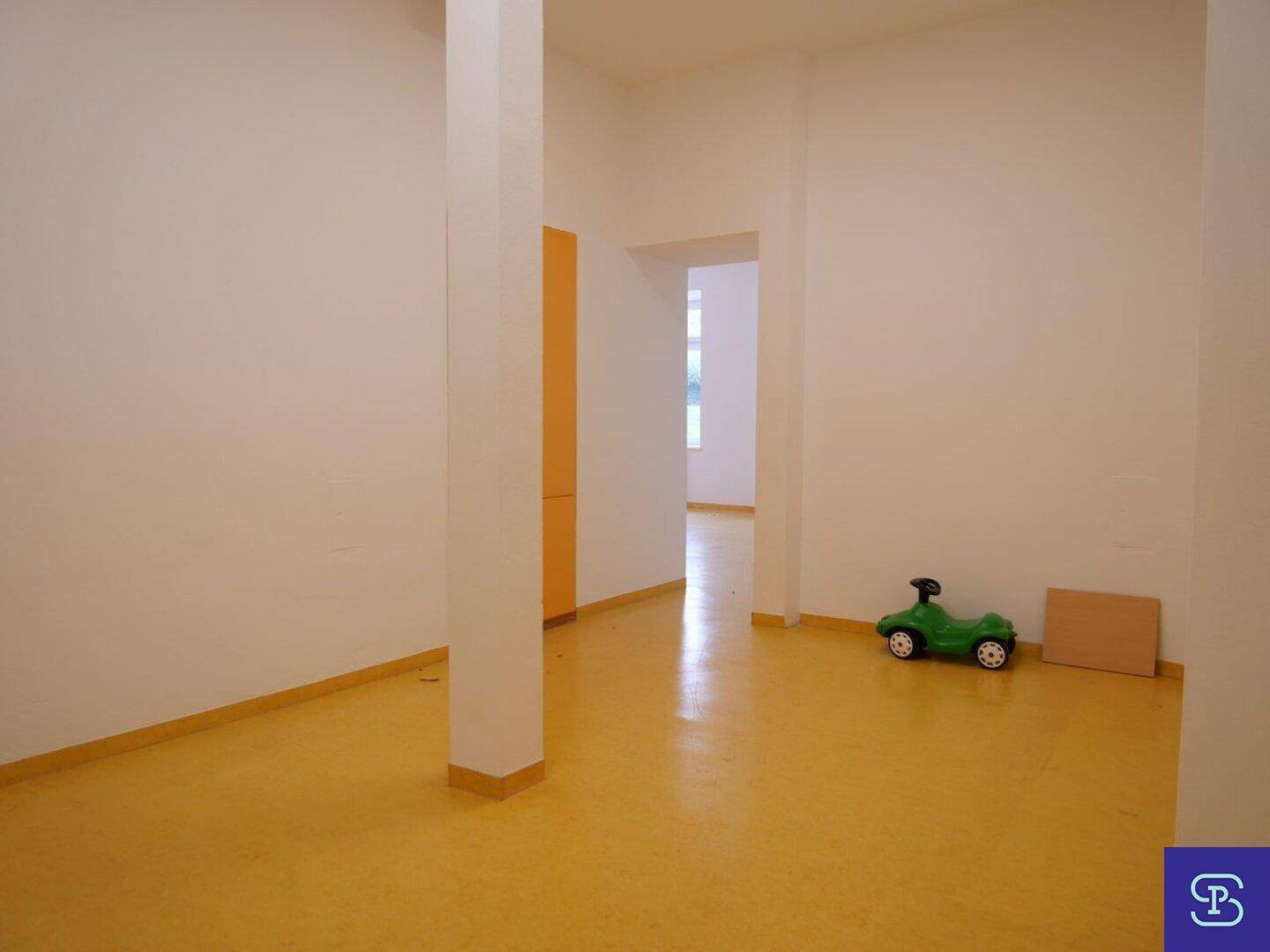 Geräumiger Raum mit gelbem Linoleumboden, weißen Wänden und einem grünen Spielzeugfahrzeug, gut beleuchtet.