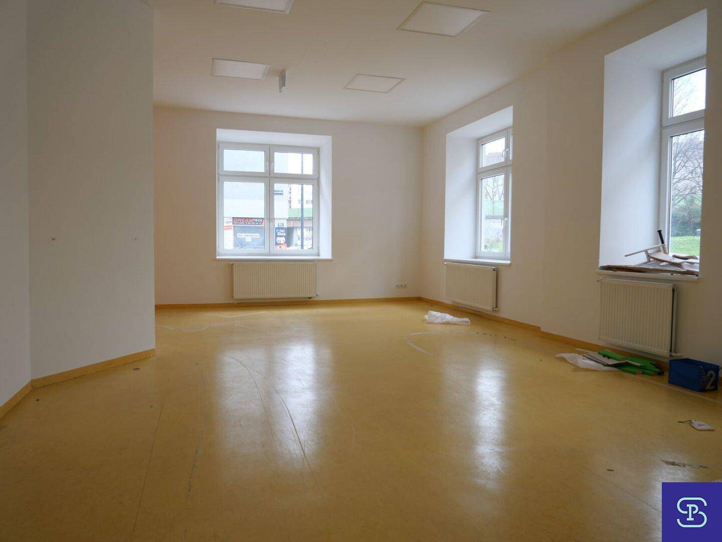 Großer, heller Raum mit gelbem Linoleumboden und mehreren Fenstern, die viel Tageslicht hereinlassen.