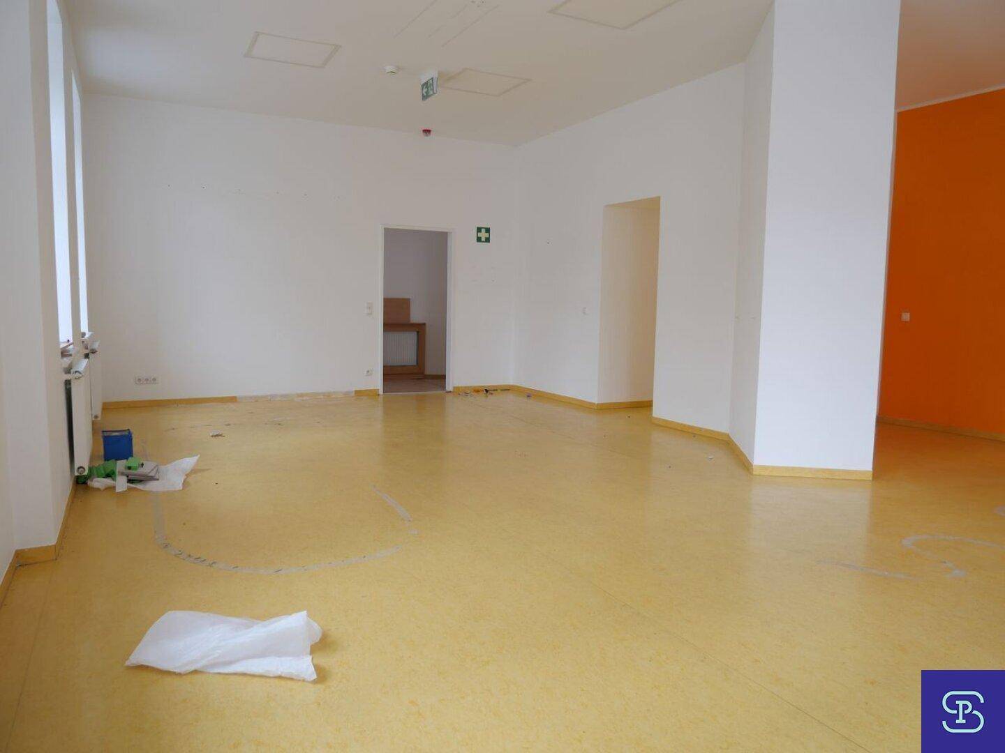 Geräumiger Raum mit gelbem Linoleumboden, weißen Wänden und einem Durchgang zu einem weiteren Bereich.