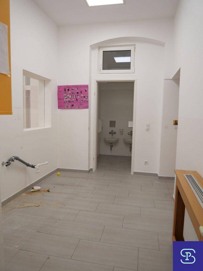 Vorbereich eines Waschraums mit hellgrauen Fliesen, weißen Wänden und einem kleinen Fenster, in gutem Zustand.