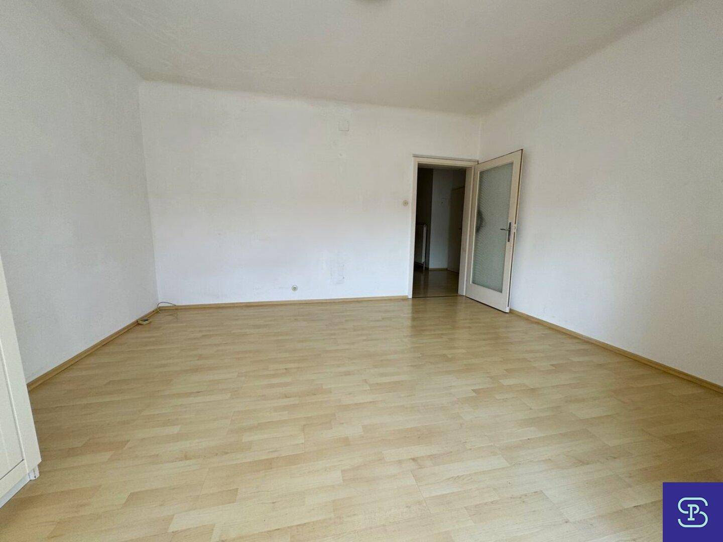 Leeres Zimmer mit hellem Laminatboden und einer Tür, die in einen Flur führt.