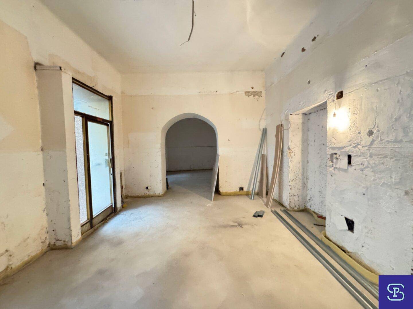 Rohbauzimmer mit Betonboden, einem großen Fenster und einem eleganten Rundbogen zu einem weiteren Bereich.