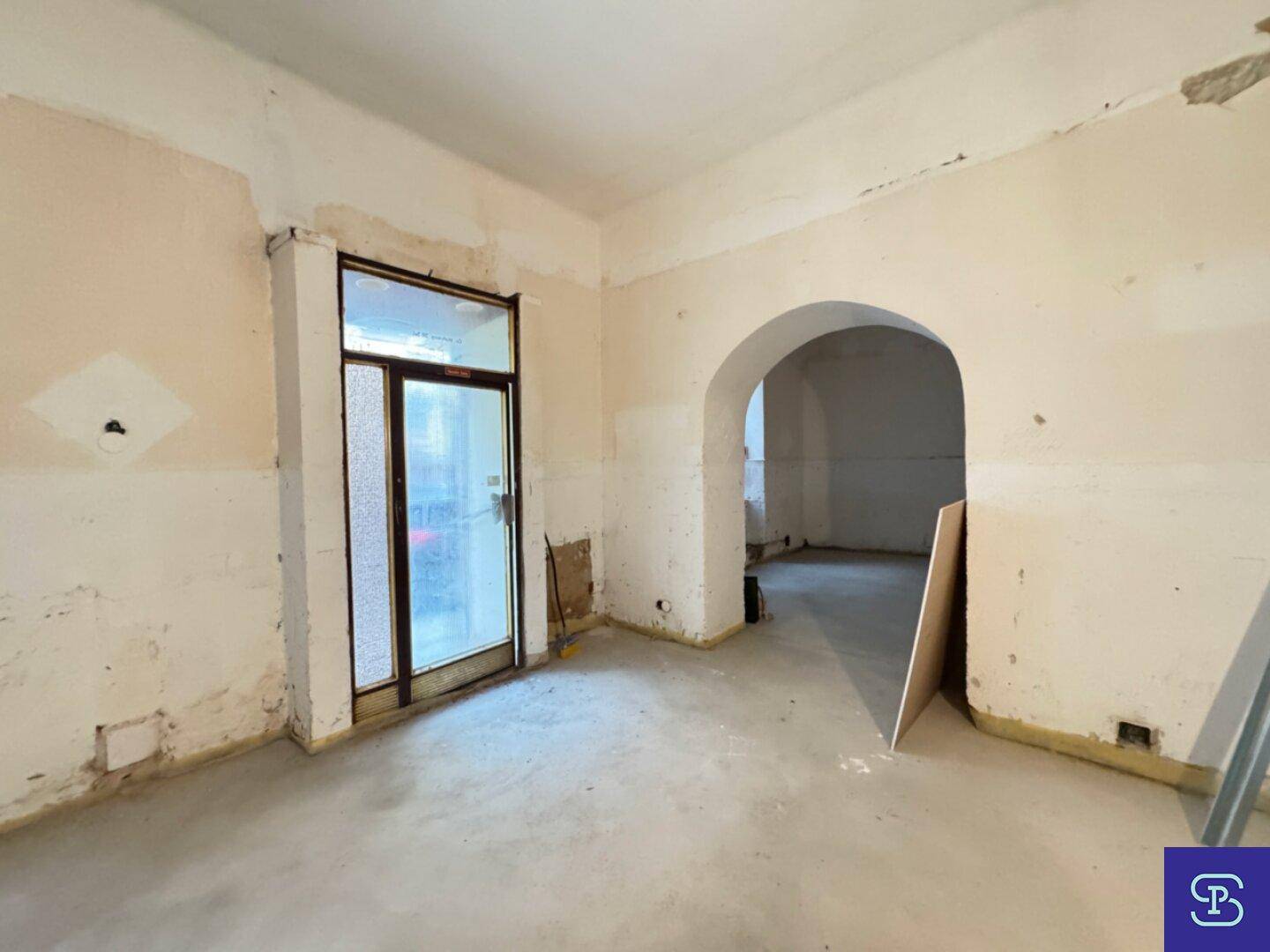 Renovierungsbedürftiger Raum mit Betonboden und einem Bogen, der zu einem weiteren Bereich führt.