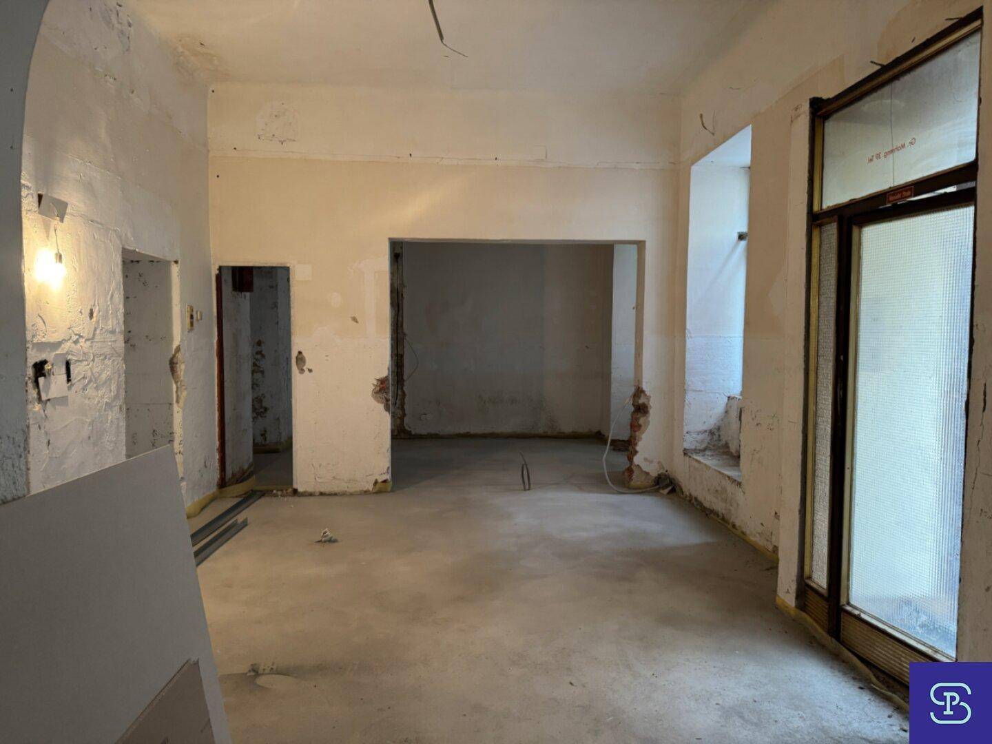 Renovierungsbedürftiger Raum mit Betonboden, freigelegten Wänden und einem Fenster, das Licht hereinlässt.