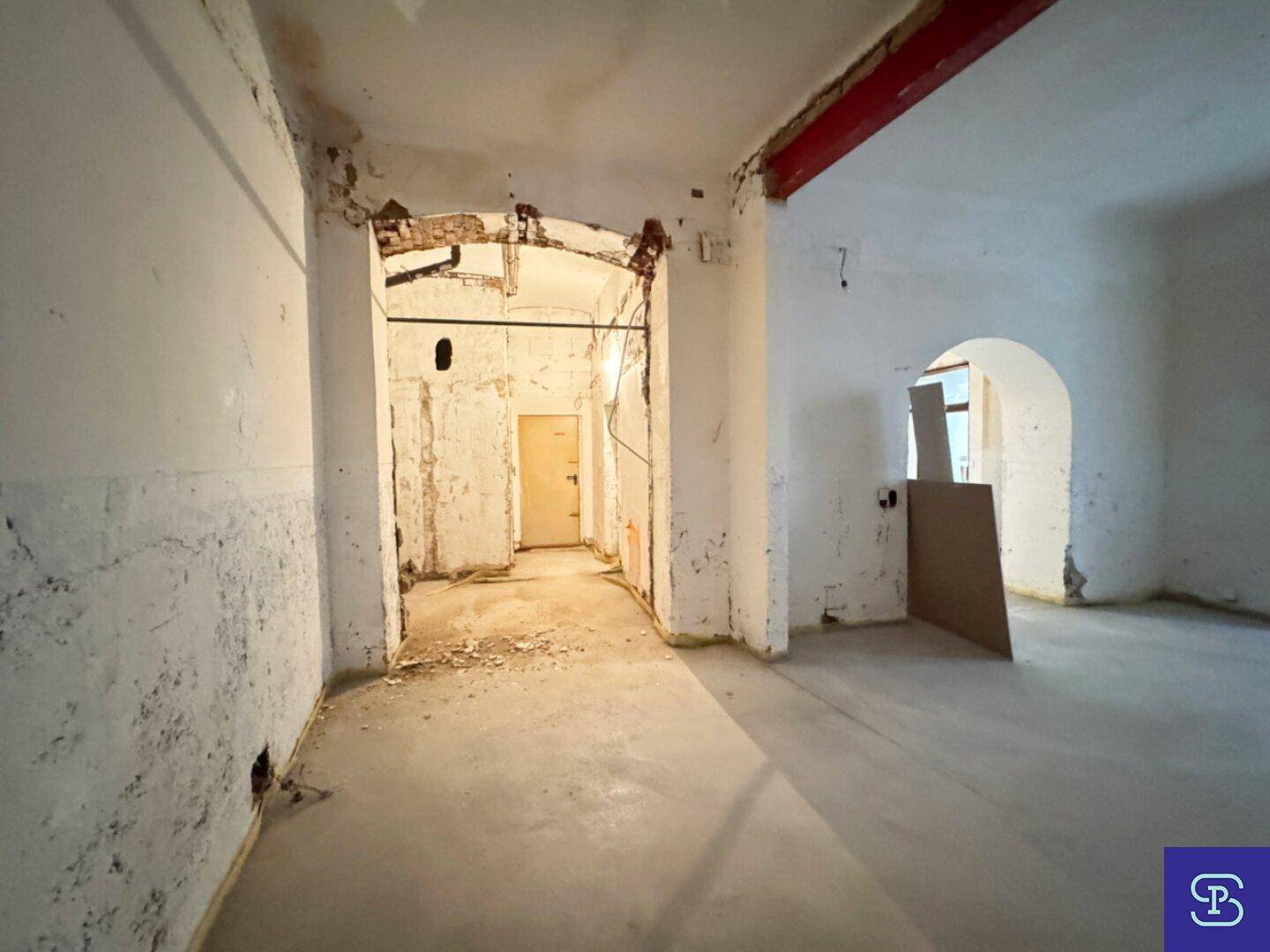 Renovierungsbedürftiger Raum mit Betonboden, freiliegendem Träger und einem Bogen im Hintergrund.