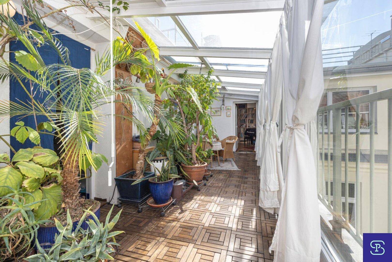 Langer Wintergarten mit Holzboden, vielen Topfpflanzen und Zugang zu weiteren Räumen.