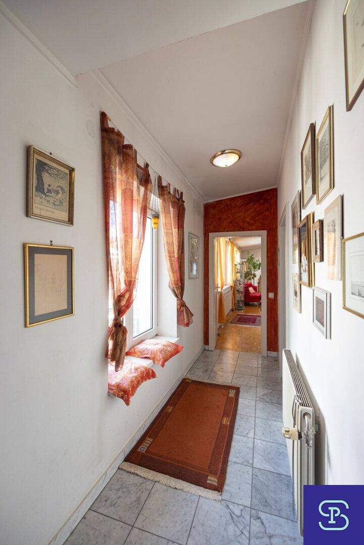 Langer Flur mit Fliesenboden, Fenster mit Vorhängen und Zugang zu weiteren Räumen.