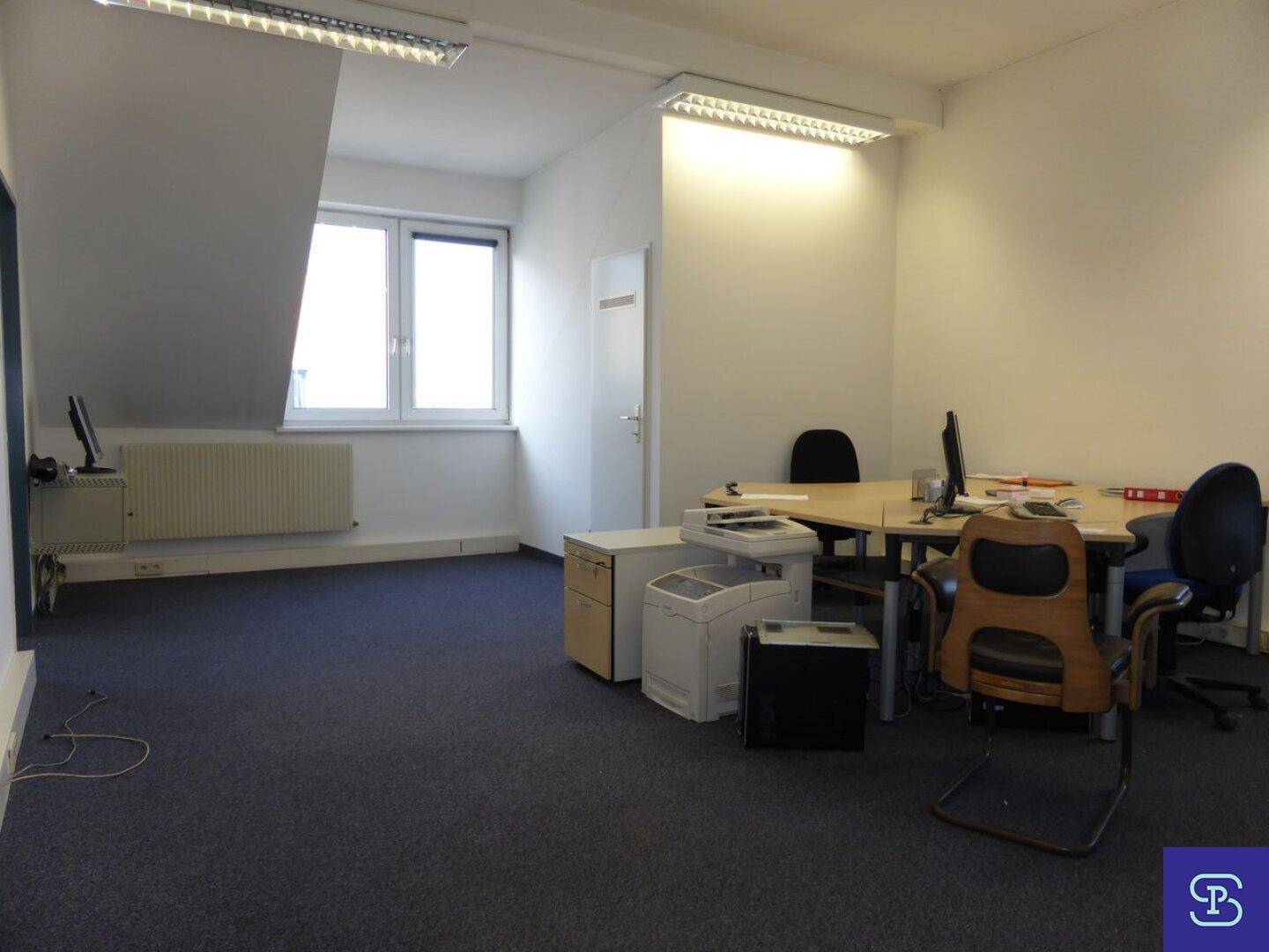 Helles Büro mit Teppichboden, Heizkörper unter dem Fenster und mehreren Arbeitsplätzen im Dachgeschoss.