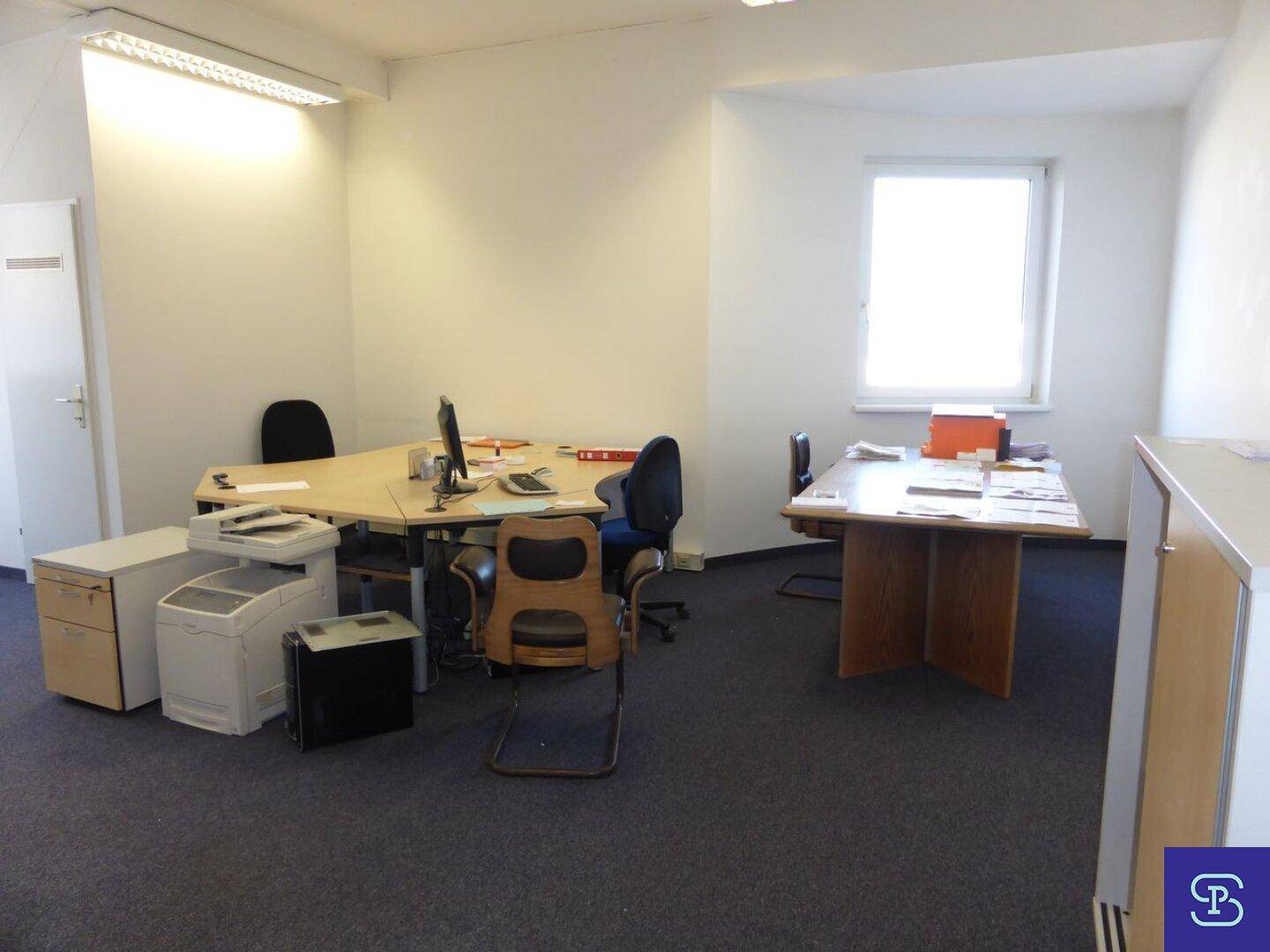 Funktionales Büro mit Teppichboden, zwei Arbeitsbereichen und natürlichem Lichteinfall durch das Fenster.