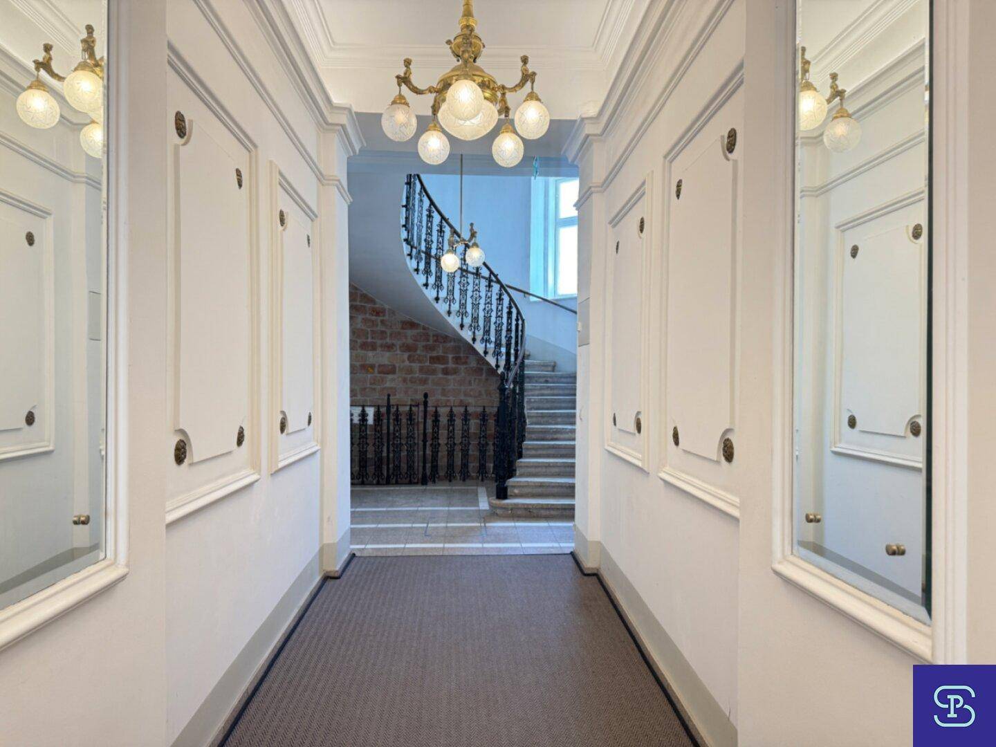 Eleganter Flur mit Stuckdecke, Kronleuchter und Spiegeln, der zu einer geschwungenen Treppe führt.