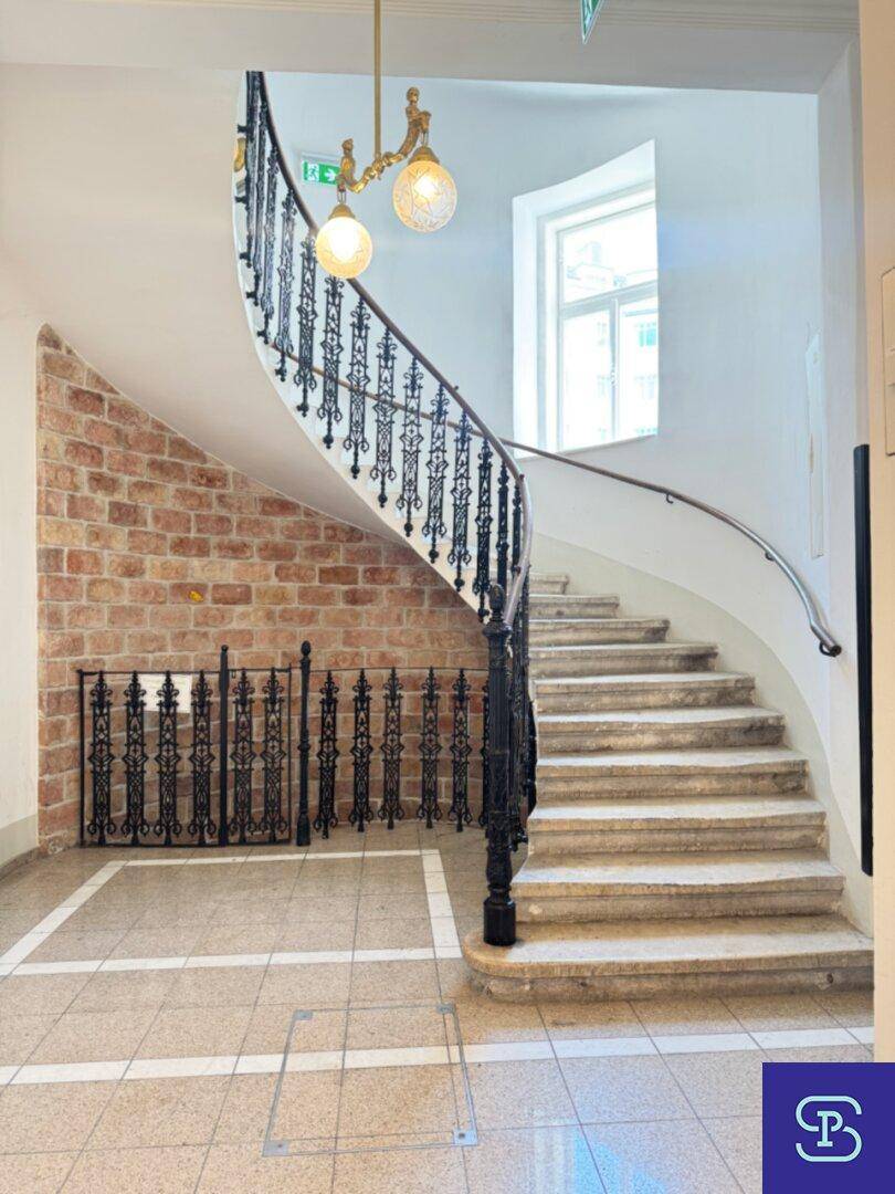 Historisches Treppenhaus mit geschwungener Steintreppe, schmiedeeisernem Geländer und freigelegter Ziegelwand.