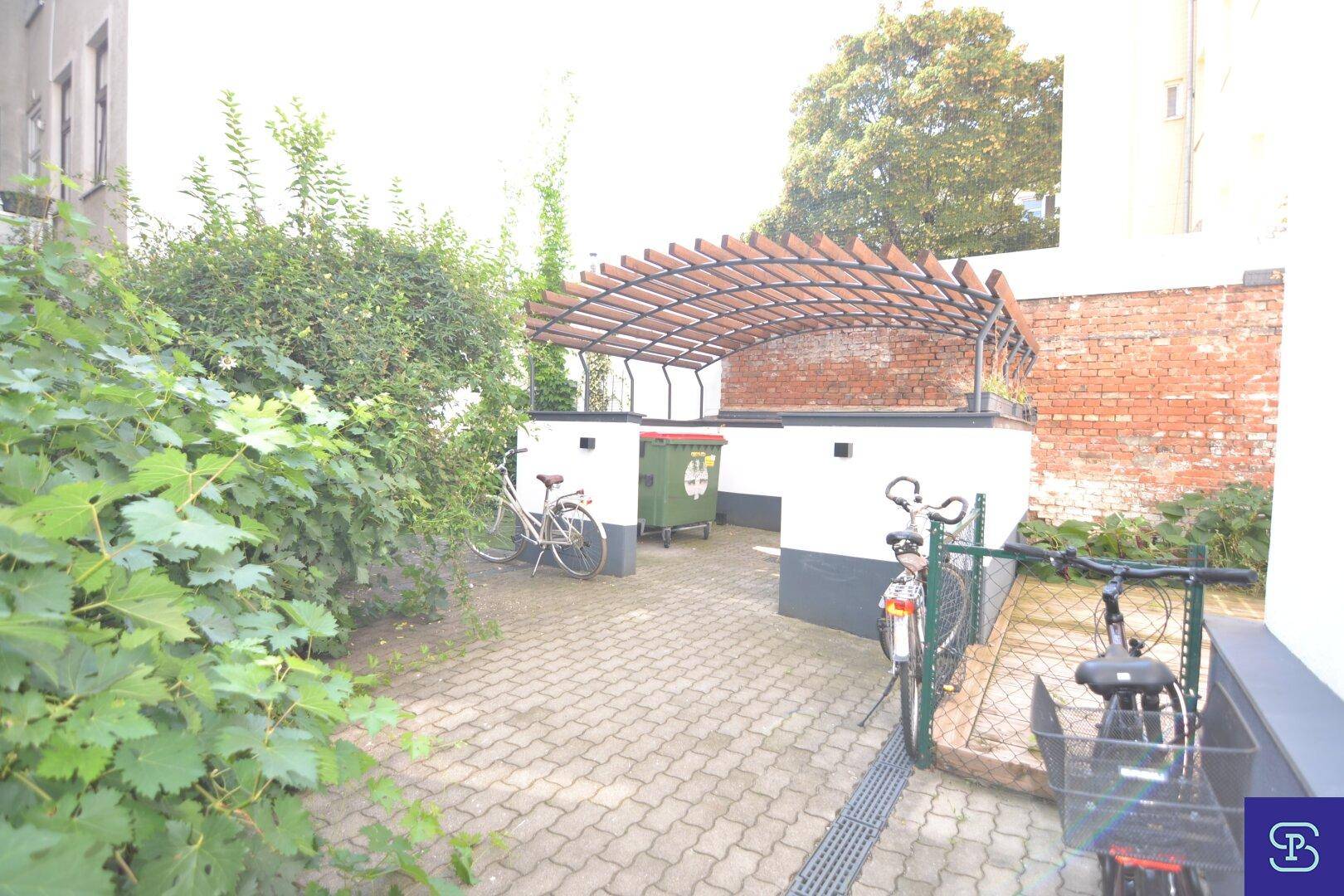 Gepflasterter Außenbereich mit Fahrradabstellplätzen, Mülltonnen und viel Grün.