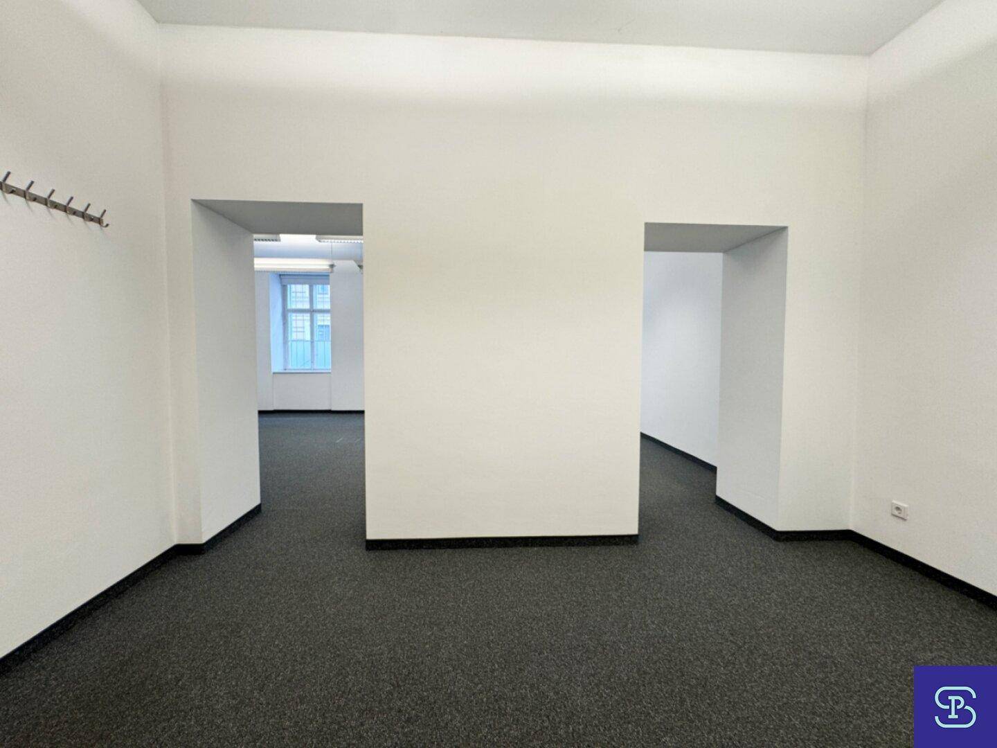 Innenansicht eines Bürobereichs mit Teppichboden und zwei offenen Durchgängen zu weiteren Räumen.