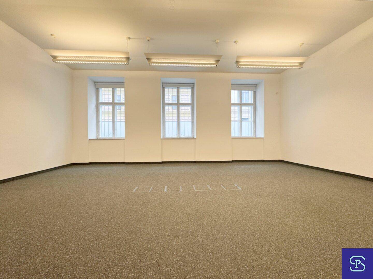 Heller Büroraum mit Teppichboden, drei Fenstern und Deckenleuchten, ideal für Arbeitsbereiche.