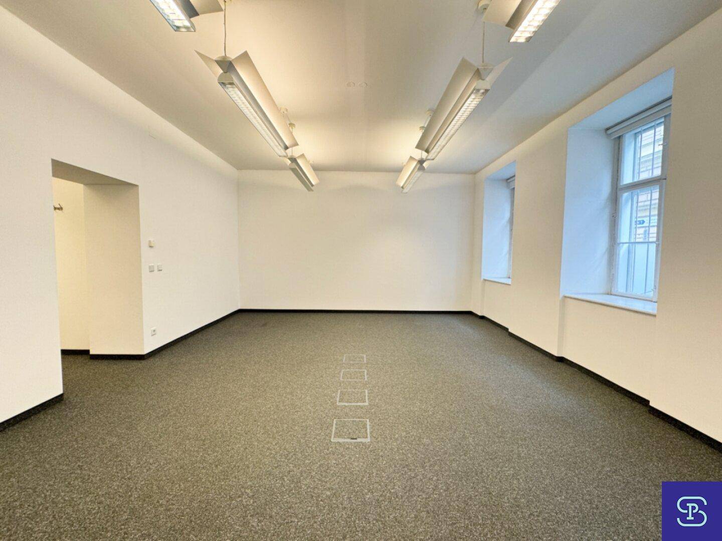 Leerer Büroraum mit Teppichboden, hohen Decken und zwei Fenstern, die für eine helle Atmosphäre sorgen.