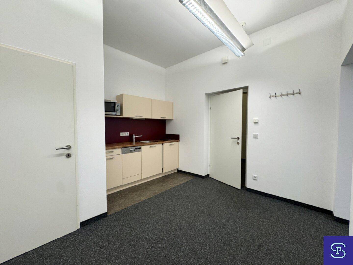 Kompakte Küchenzeile in einem Büroraum mit Spüle, Mikrowelle und Hängeschränken.