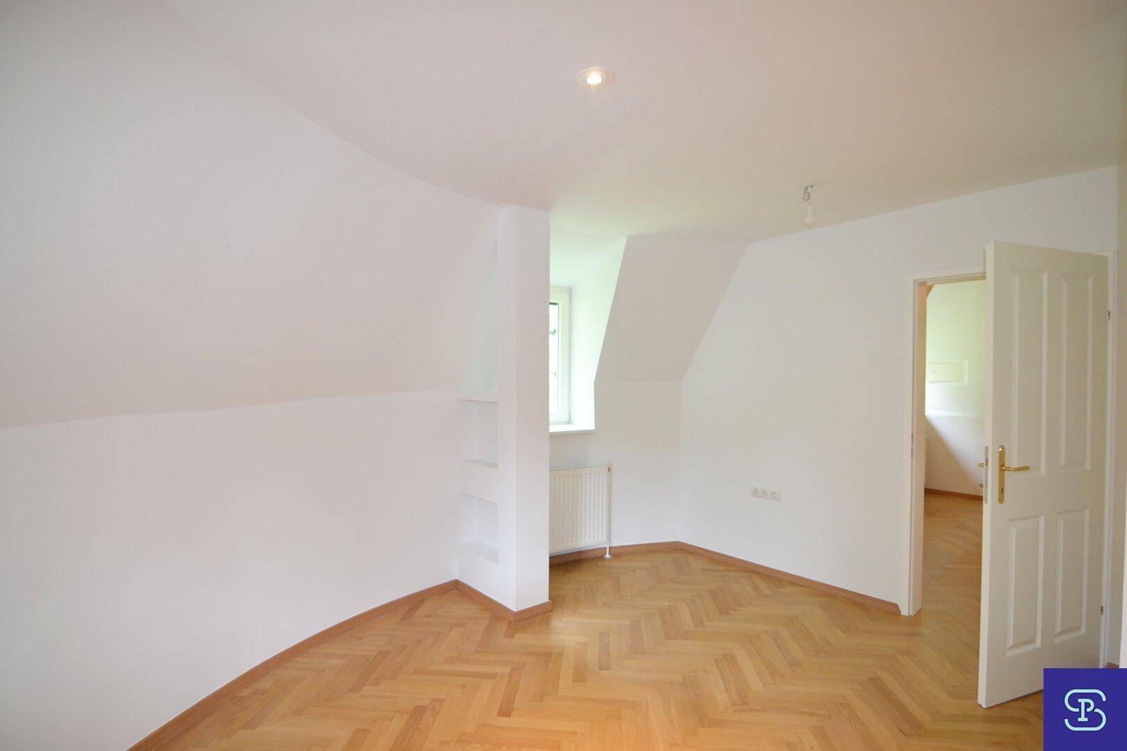 Dachgeschosszimmer mit Fischgrätparkett, Fenster und offener Tür zu einem weiteren hellen Raum.