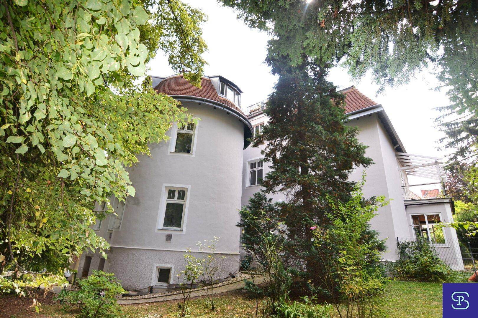 Historisches Gebäude mit markanter runder Fassade, roten Dächern und umgeben von viel Grün.