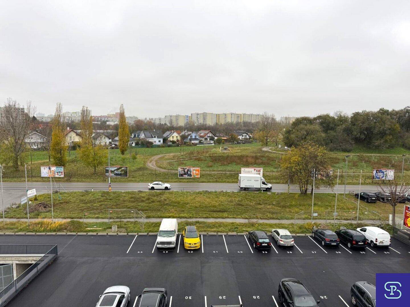 Blick von oben auf einen Parkplatz mit Fahrzeugen, einer Straße und Wohngebäuden im Hintergrund.