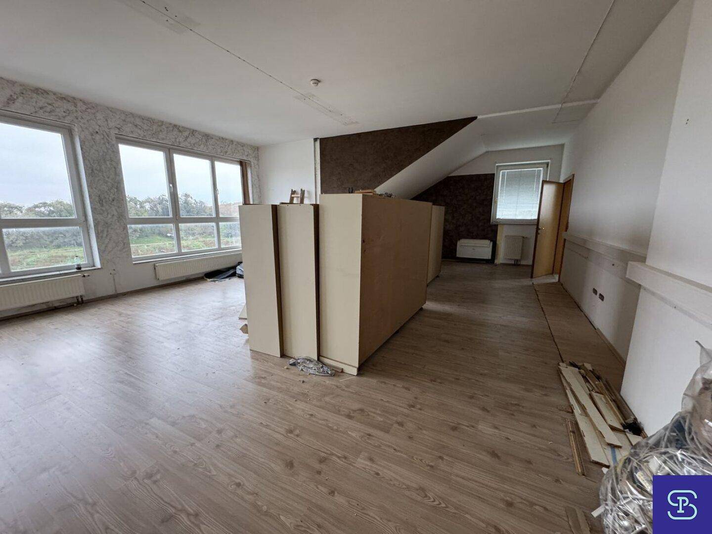 Weitläufiger Raum mit Laminatboden, großen Fenstern und schräger Decke, der auf eine bevorstehende Renovierung wartet.