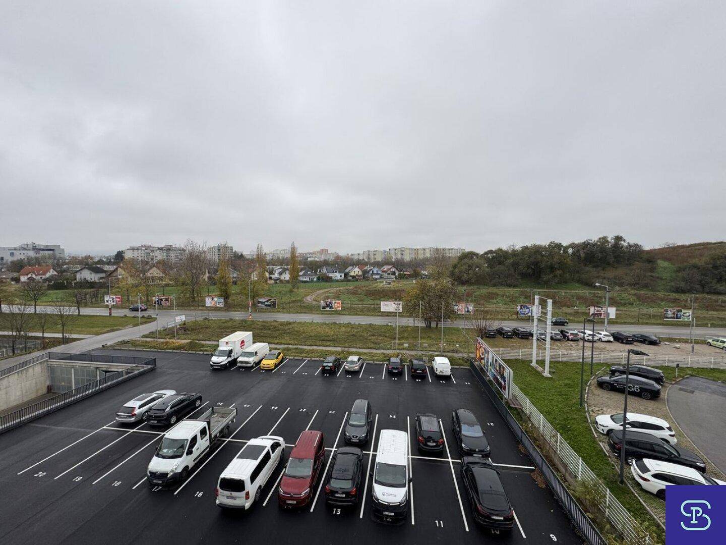 Großer Außenparkplatz mit zahlreichen Fahrzeugen und Blick auf die umliegende Landschaft und Wohnsiedlung.