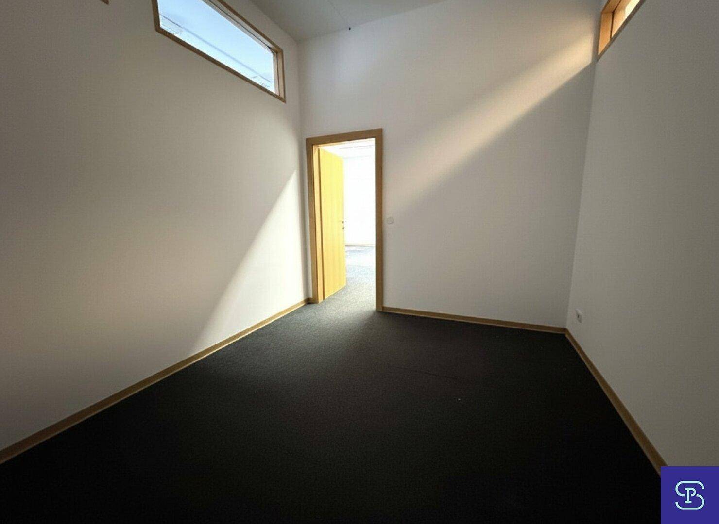 Leerer Büroraum mit Teppichboden, weißen Wänden und einer offenen Tür, die in einen weiteren Bereich führt.
