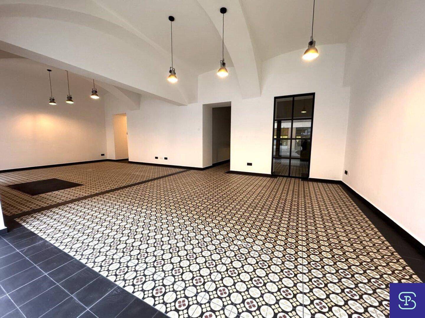 Großer, leerer Gewerberaum mit hohen Decken, großen Fenstern und gemustertem Fliesenboden.