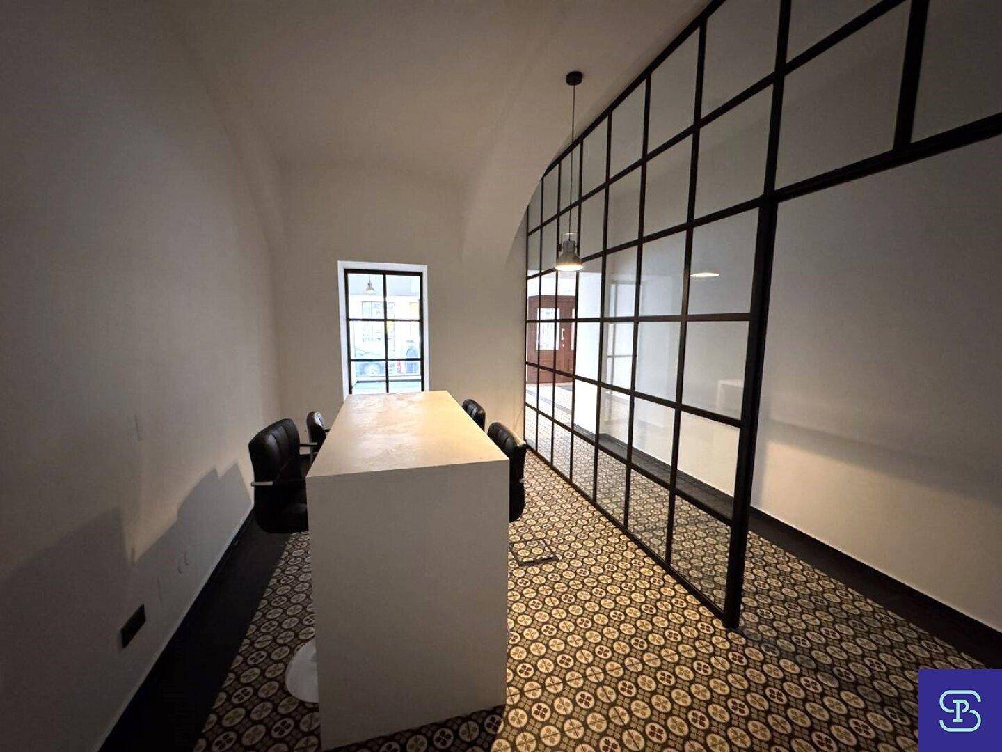 Heller Büroraum mit Glaswänden, einem großen Tisch und gemusterten Bodenfliesen.