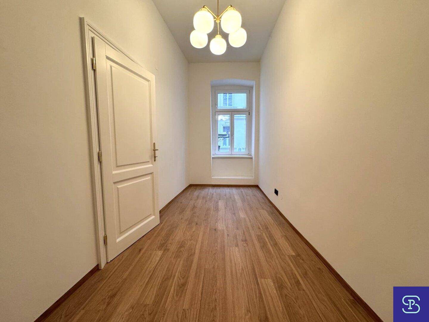 Leerer Raum mit Holzboden, weiß gestrichenen Wänden und einem Fenster zur Straße.