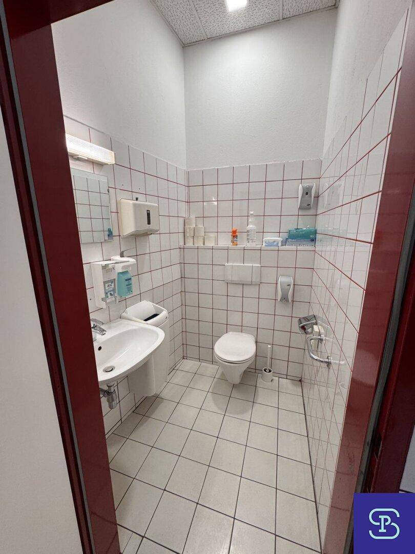 Saubere Toilette mit Waschbecken und Wandfliesen, ausgestattet mit grundlegenden Hygieneartikeln und einem Spiegel.