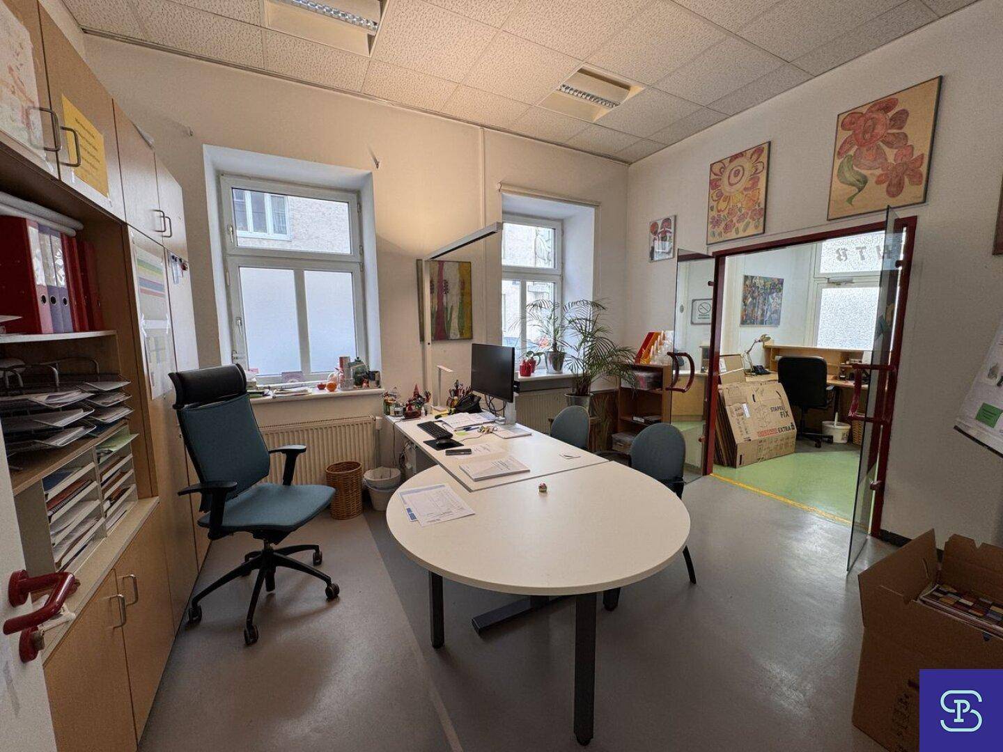 Büro mit hellem Boden, Schreibtisch und Besprechungstisch, mit Fenstern und Zugang zu weiteren Räumen.