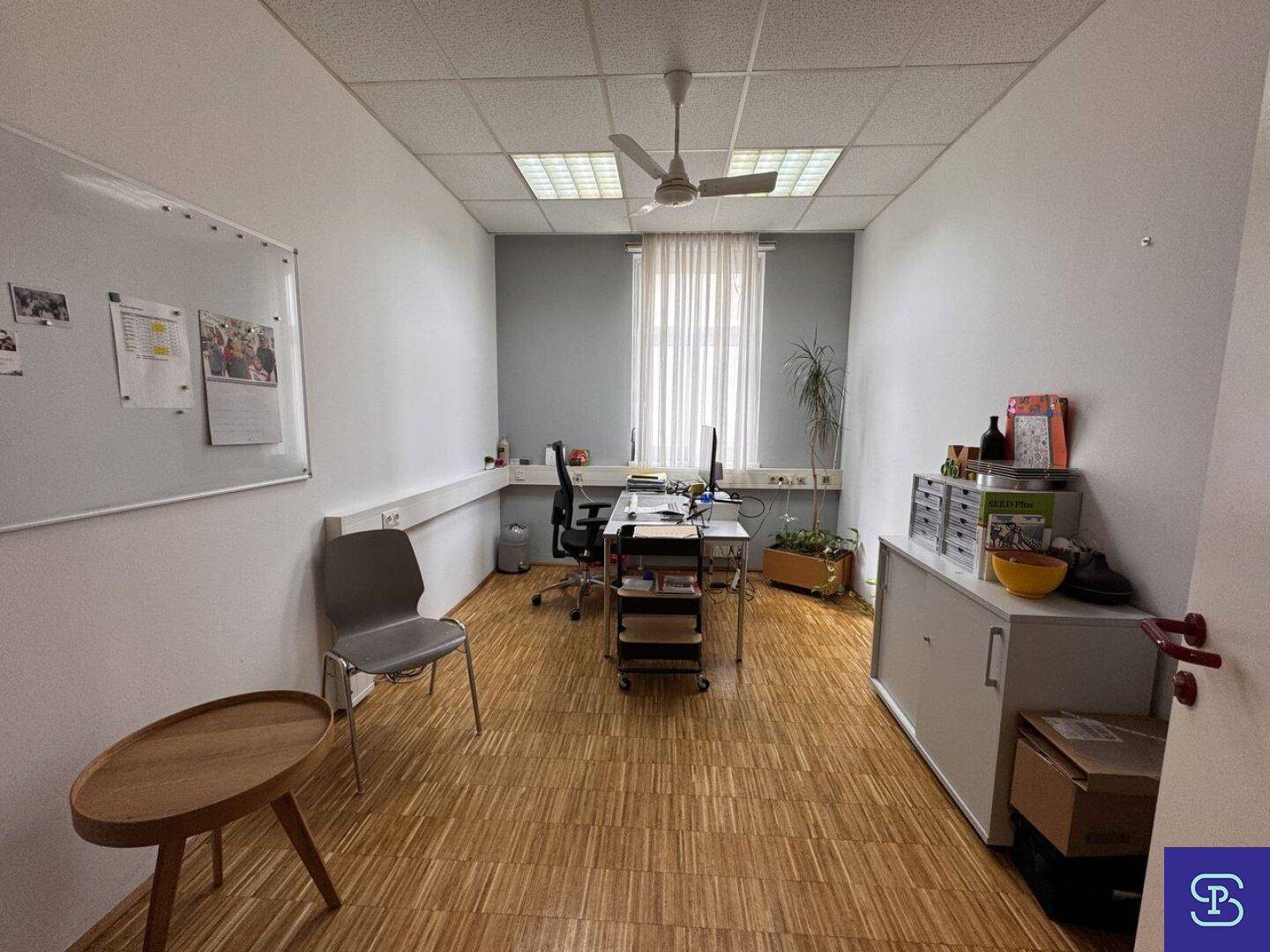 Büro mit Holzparkett, Deckenventilator und einem Arbeitsplatz, umgeben von hellen Wänden und einem Fenster.