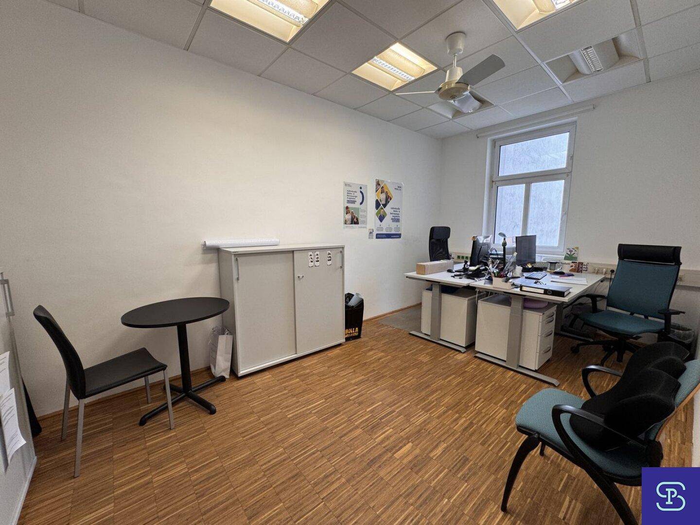 Büro mit Holzparkett, Deckenventilator und zwei Arbeitsplätzen, ausgestattet mit einem kleinen Besprechungstisch.