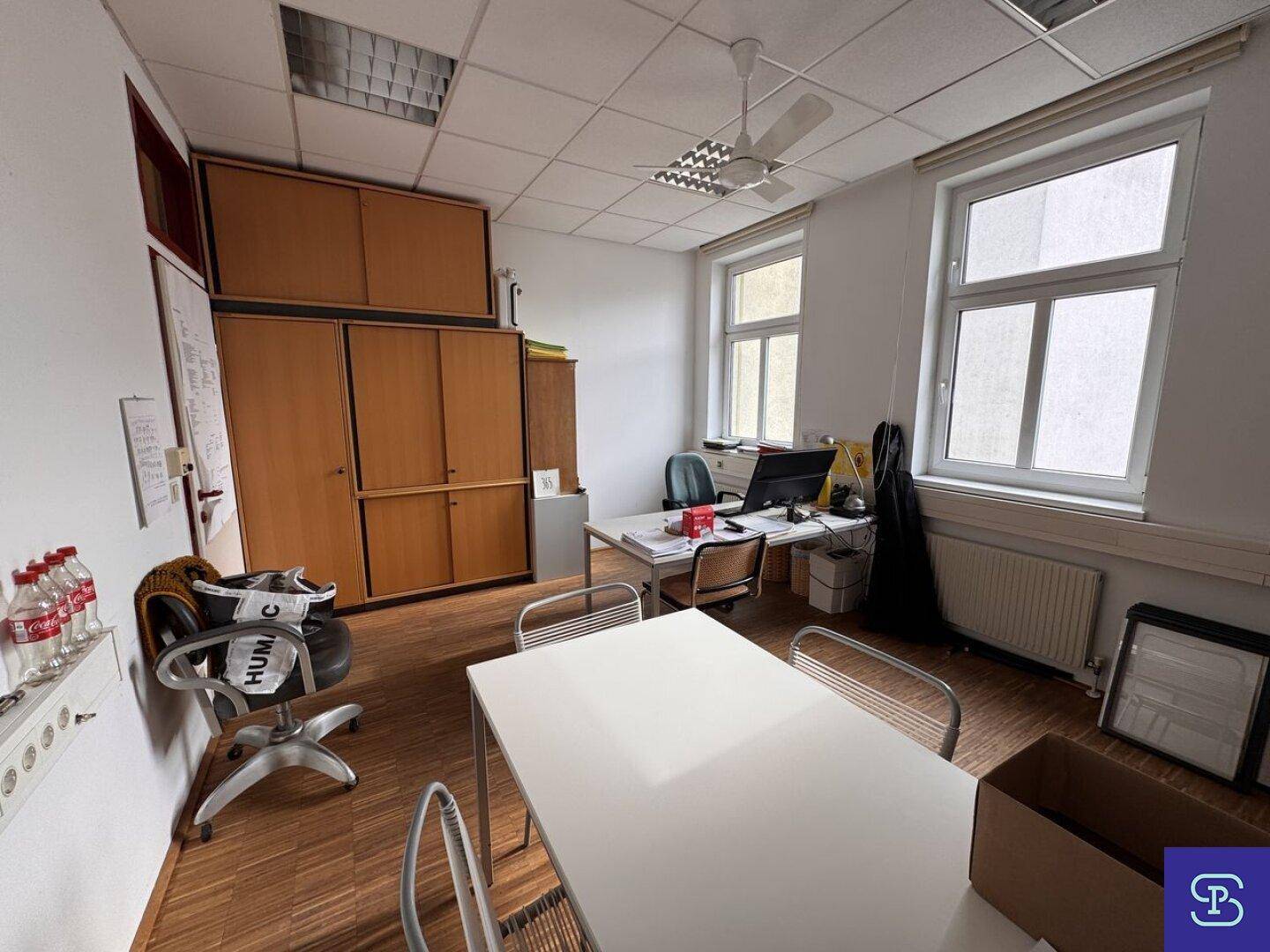 Büro mit Holzparkett, Deckenventilator und einem Arbeitsplatz, umgeben von Holzschränken und großen Fenstern.