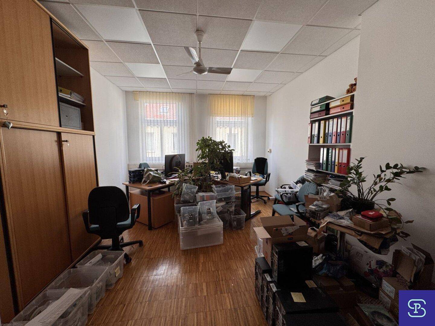 Büro mit Holzparkett, Deckenventilator und mehreren Arbeitsplätzen, mit viel Stauraum und zwei Fenstern.